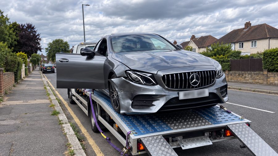 A grey Mercedes-Benz with front-end damage strapped onto a flatbed transporter